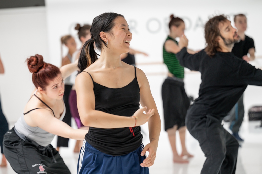 A dancer smiles in mid-practice with other dancers behind her during a training session in a dance studio